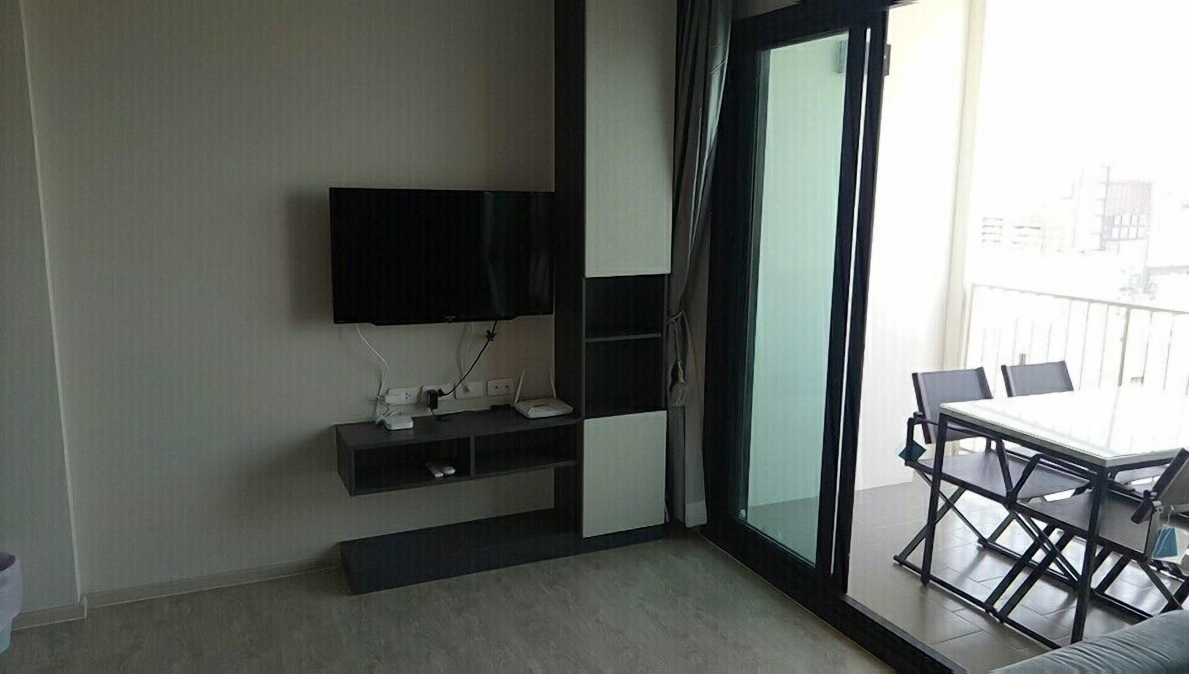 Una sala moderna con un televisor de pantalla plana montado en la pared, una pequeña consola con dispositivos electrónicos, y un balcón con una mesa y sillas de comedor.