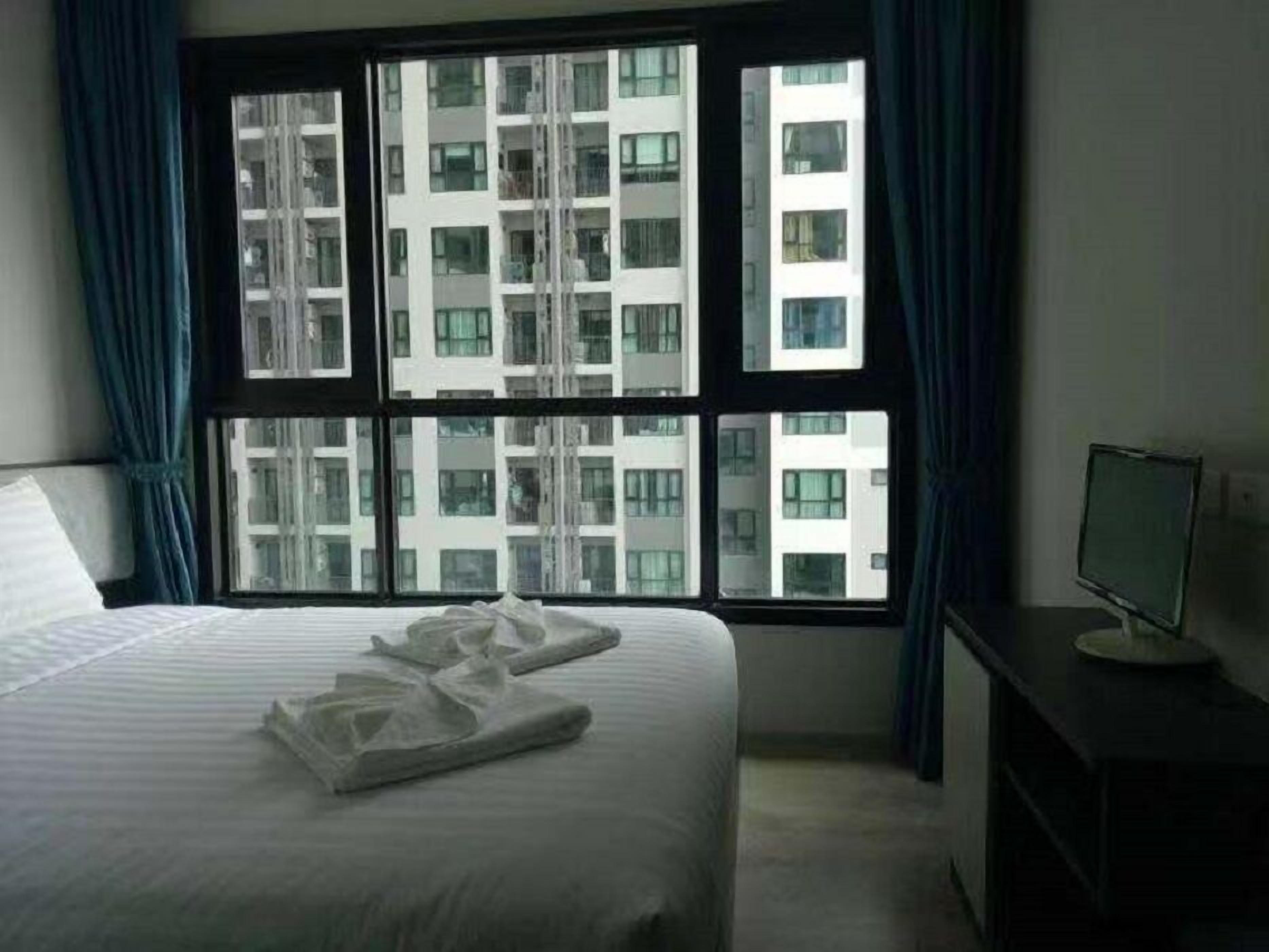 A hotel room with a large bed, a desk with a computer, and a view of a high-rise building through the window.