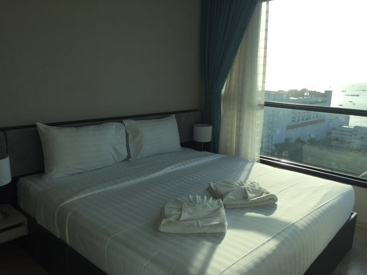 A hotel room with a large bed, a nightstand, a lamp, and a window with a view of buildings and water.