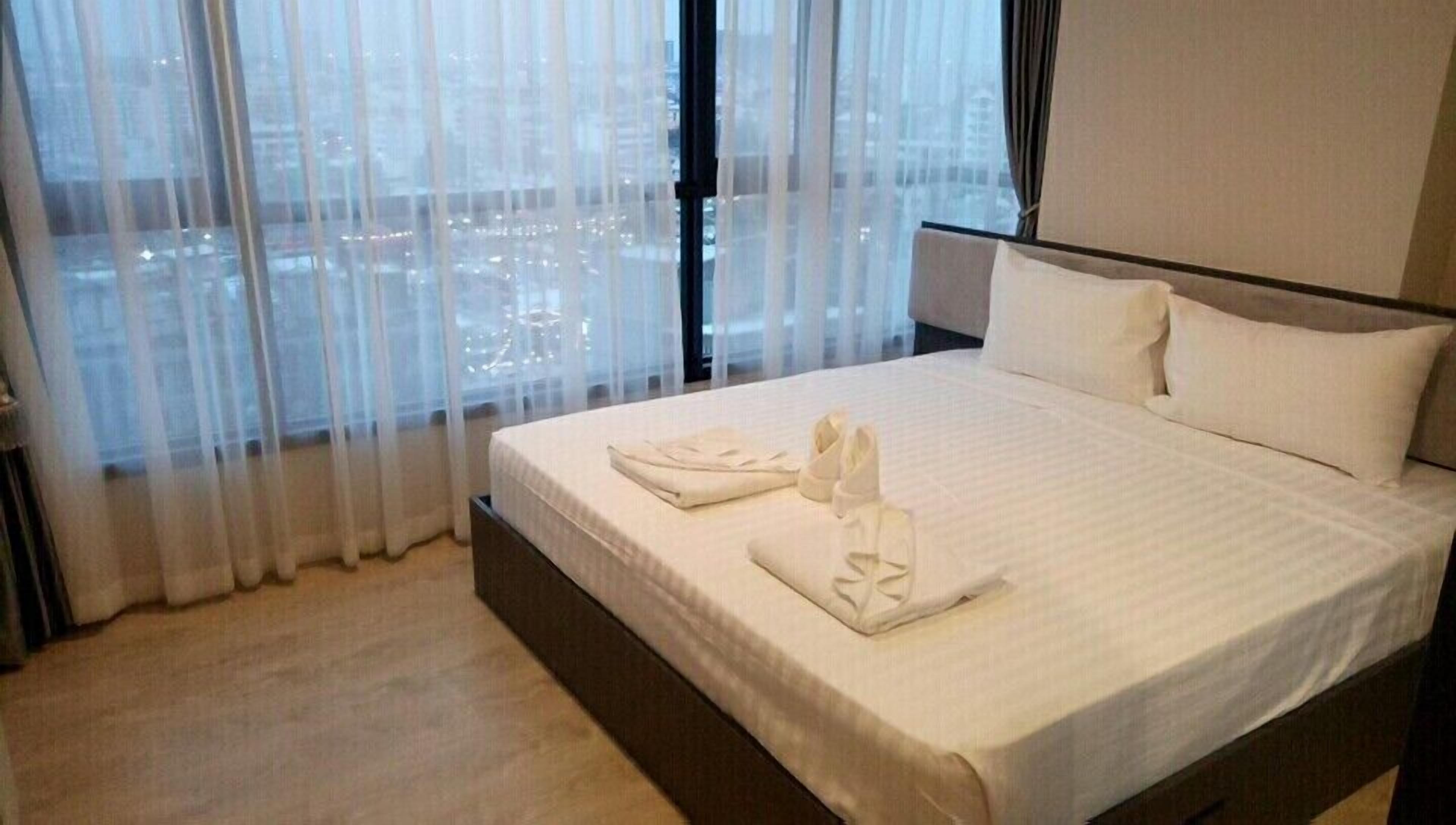 A hotel room with a large bed, white linens, and a view of a cityscape through sheer curtains.