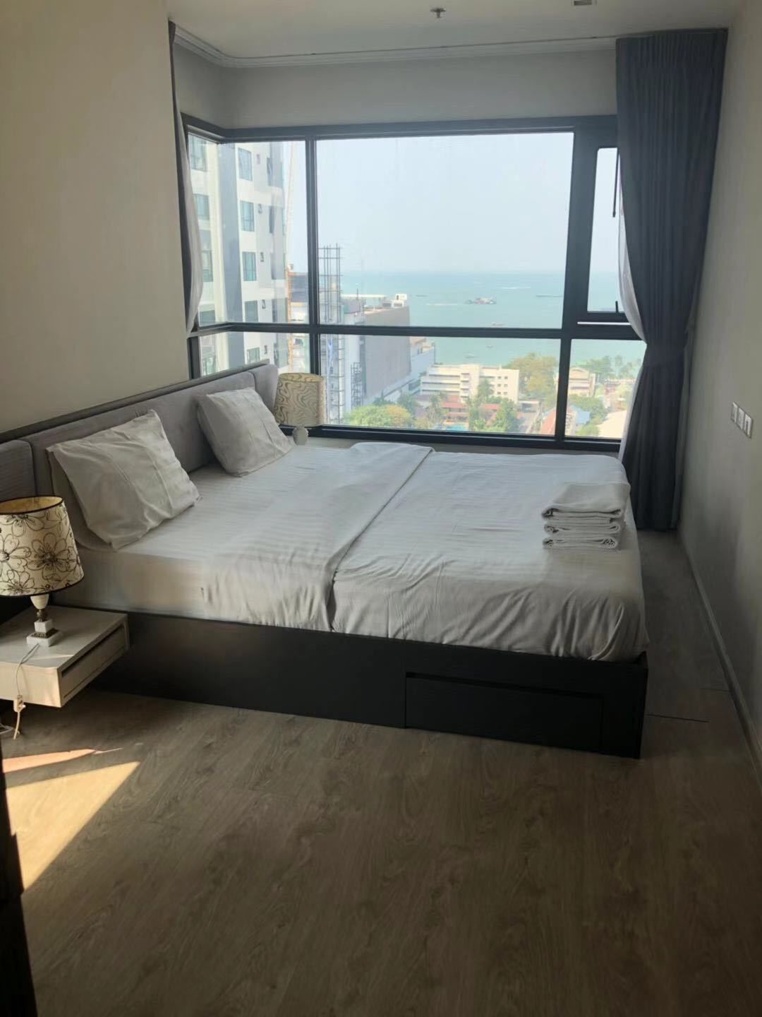 A modern bedroom with a large bed, a nightstand, a lamp, and a view of the sea through a large window.