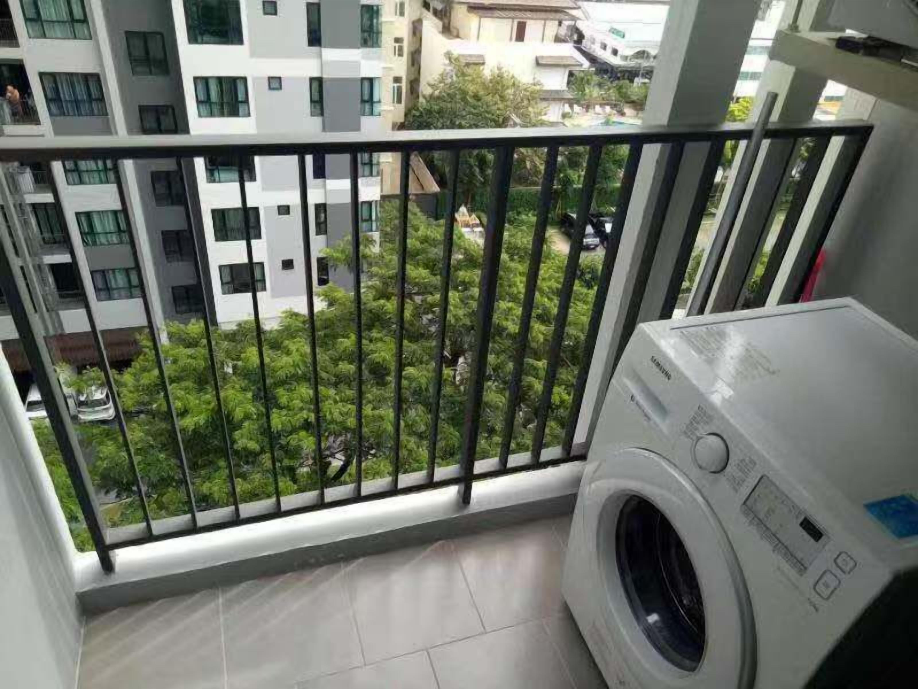 A balcony with a washing machine, a view of a residential area with greenery and buildings.
