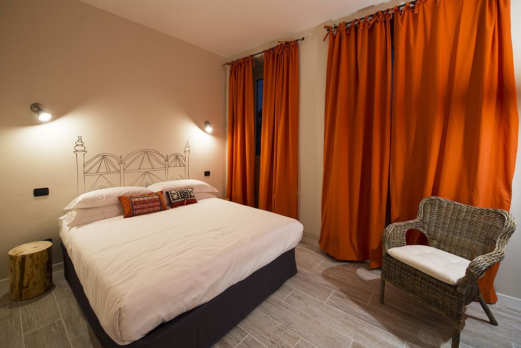 Een slaapkamer met een bed, oranje gordijnen, een rieten stoel en een klein houten krukje.
