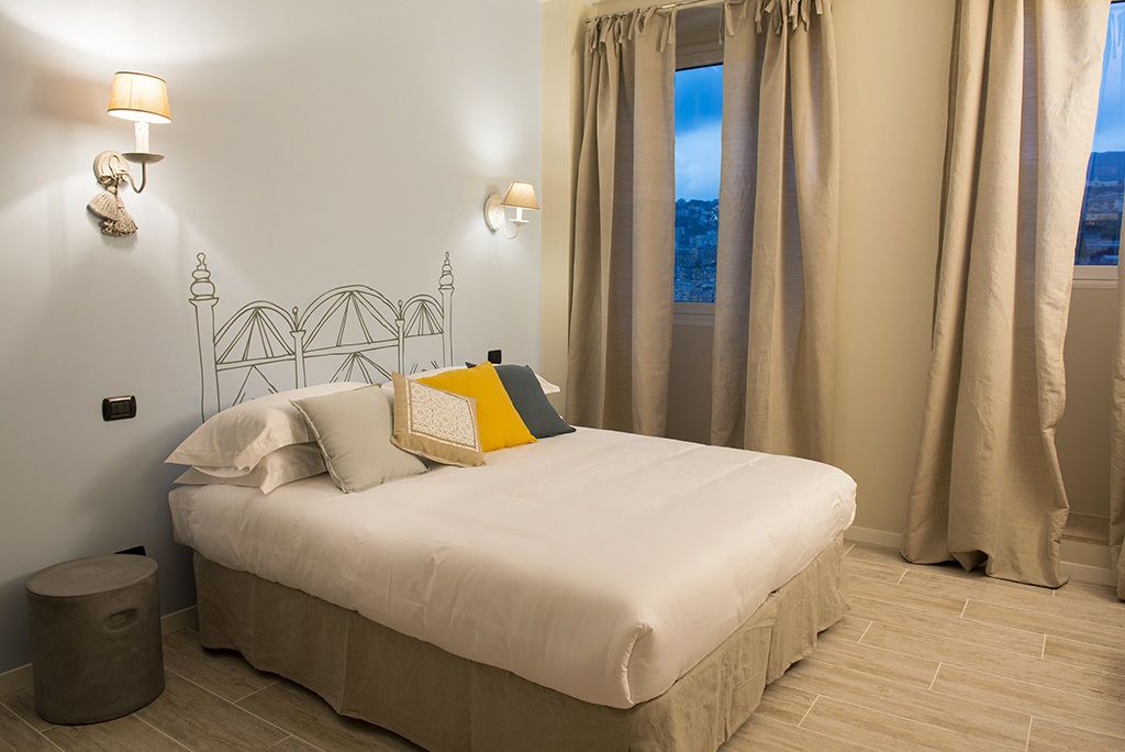 Cama bem arrumada com edredom branco e almofadas decorativas, uma luminária na parede e vista para o exterior através de uma janela com cortinas.