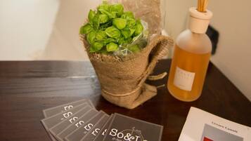 A room with a bed, a table with a bottle of reed diffuser, a basket of green leaves, and a booklet titled "S&O Leo".