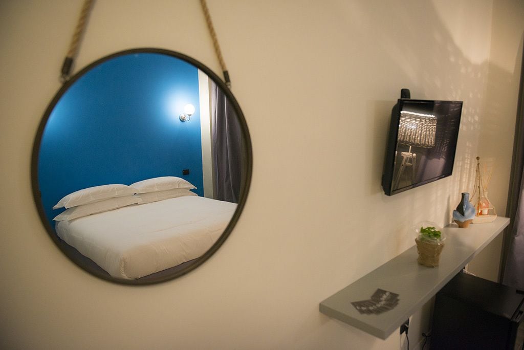 A room with a bed, a round mirror, a flat-screen TV, and a small shelf with decorative items.