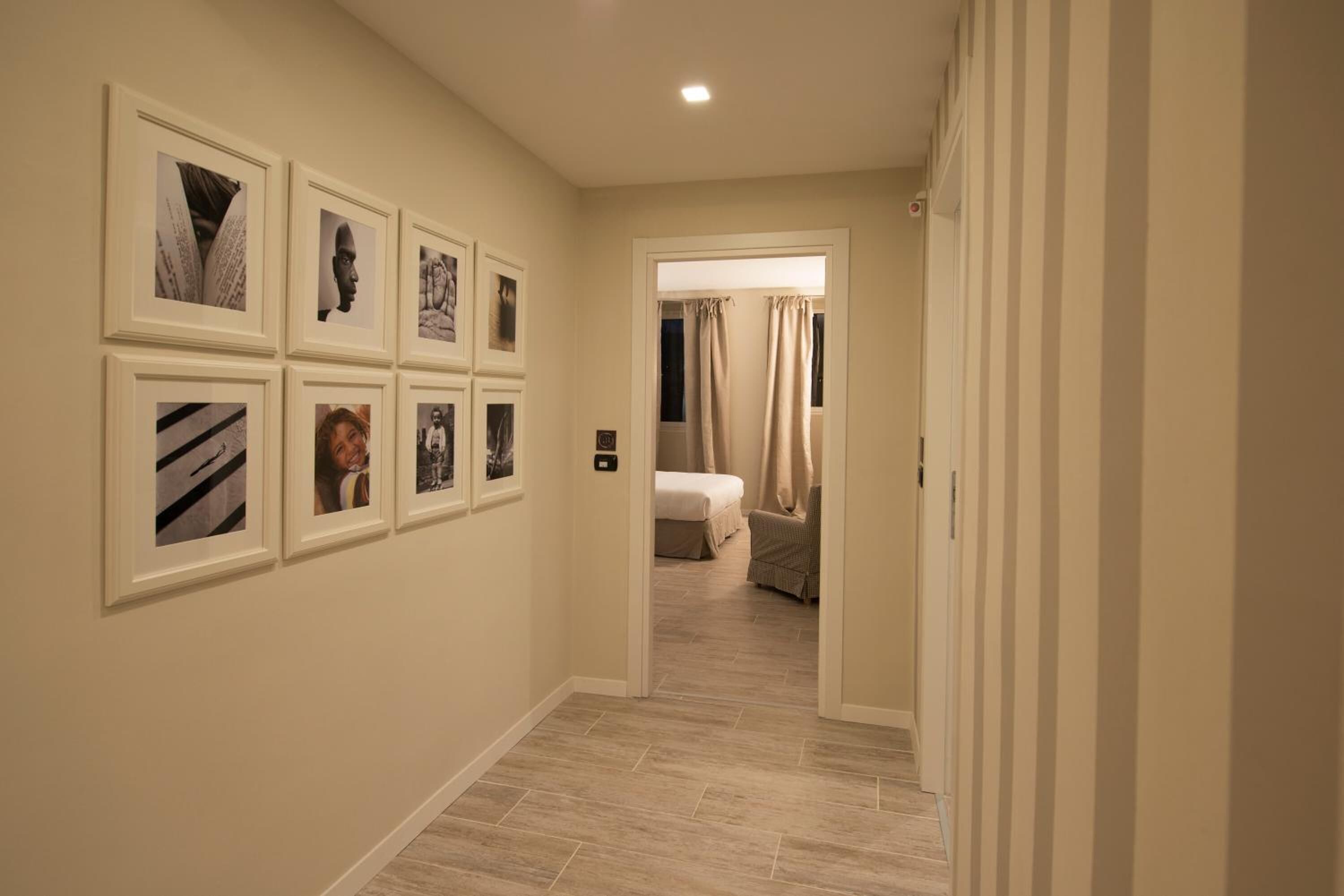 A hallway with framed pictures on the wall, a bed in the background, and a door leading to another room.