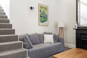 Un salon avec un canapé gris, une table basse en bois, un escalier recouvert d’une moquette à motifs, une lampe et un tableau encadré représentant Melbourne.