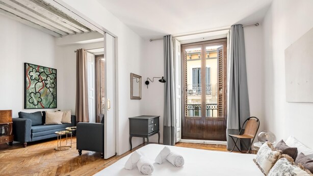 Hotellihuone, jossa on puinen sänky, sininen sohva, pieni työpöytä ja parveke, jolta on näkymä rakennuksiin.