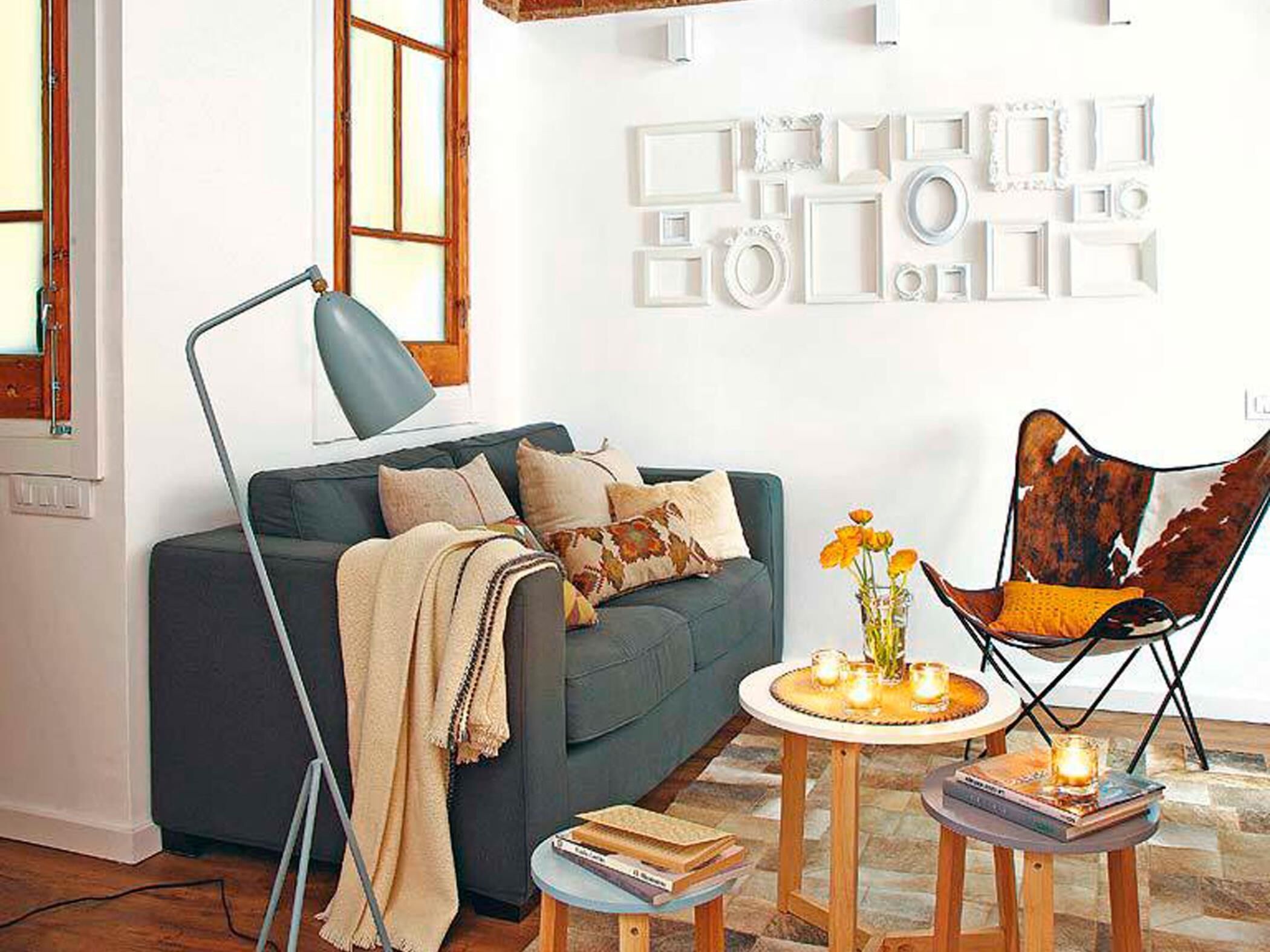Un salon comprenant un canapé, un fauteuil, une lampe, une petite table avec des livres et des bougies, et un mur orné de cadres décoratifs.