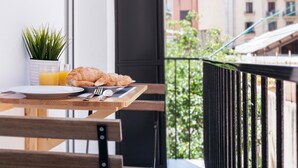 Un balcón con una mesa de madera dispuesta para el desayuno, incluyendo croissants, un plato, un tenedor y un vaso de zumo de naranja. Hay una planta en maceta sobre la mesa, y se aprecia una vista con edificios y vegetación.