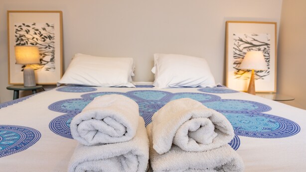 Ein ordentlich bezogenes Bett mit einer blau-weißen Tagesdecke, zwei weißen Kissen und zwei gerollten weißen Handtüchern.