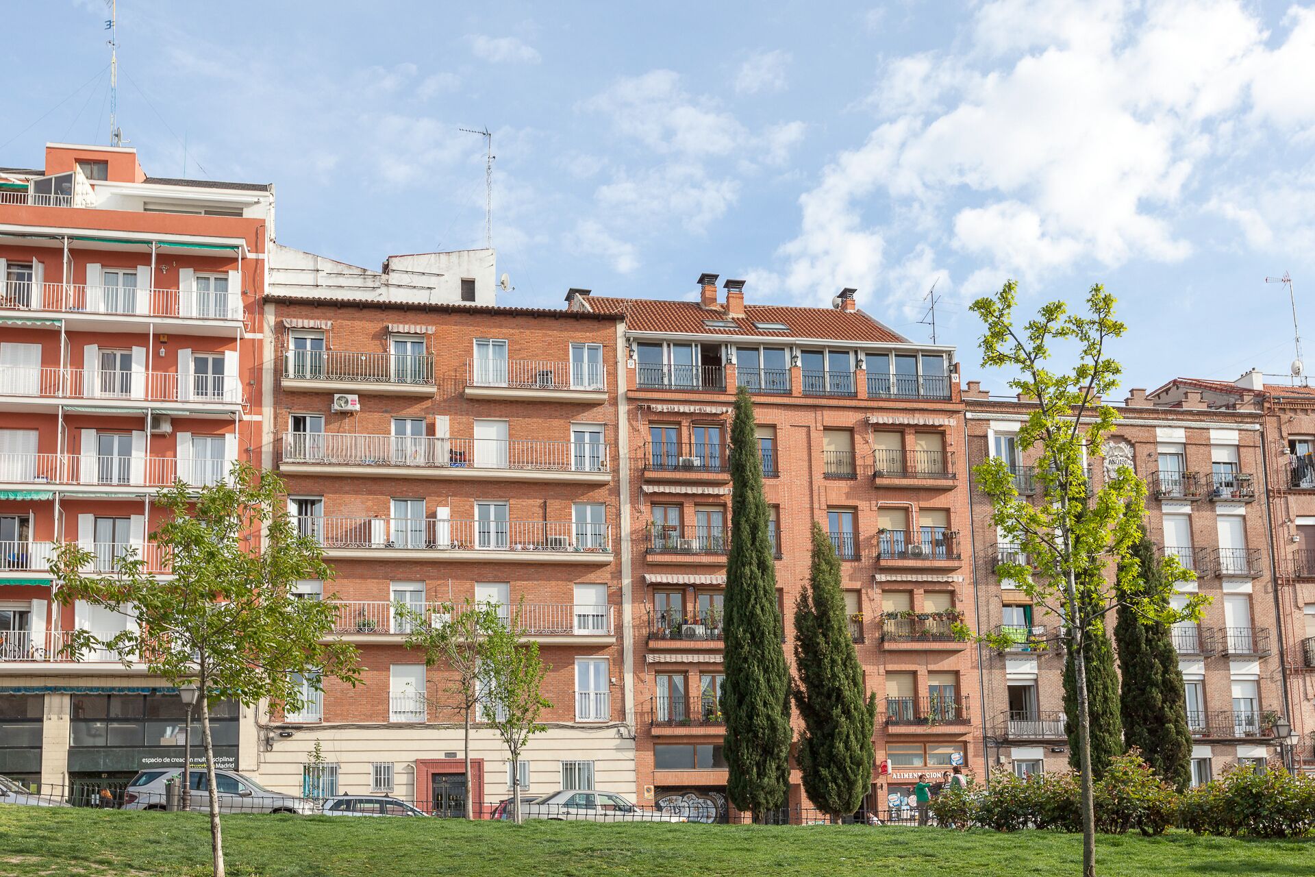 A multi-story residential building with balconies, a grassy area in front, and parked cars.