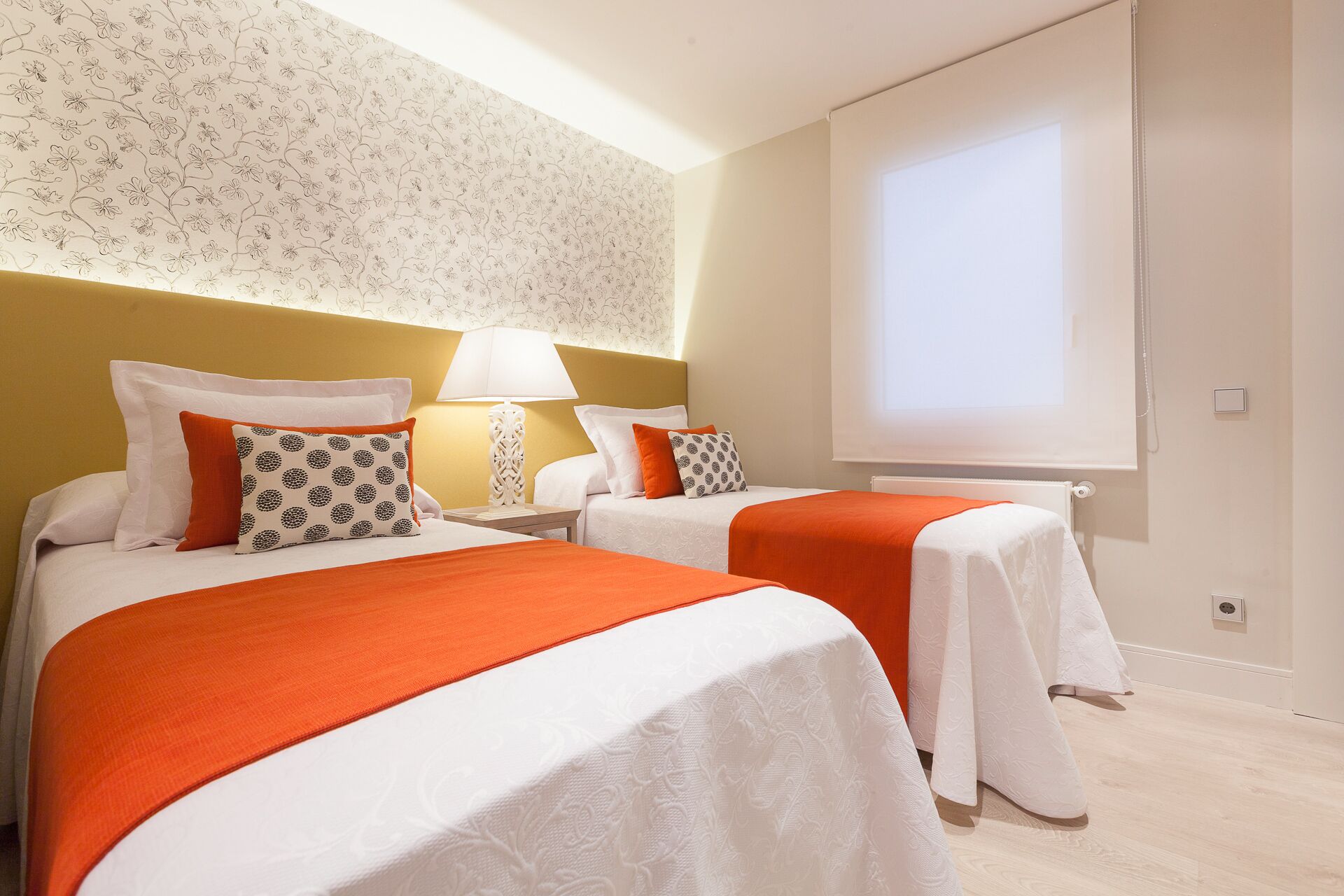 Hotelkamer met twee bedden, elk voorzien van witte beddengoed en oranje accenten, behang met een patroon, een witte lamp en een scherm voor een beamer aan de muur.