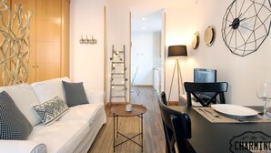 A modern living room with a white sofa, a coffee table, and a dining area with a table set for two. There is a ladder leaning against the wall and a lamp on a tripod. - Huertas studio - Apartment in the center of Madrid (Madrid)
