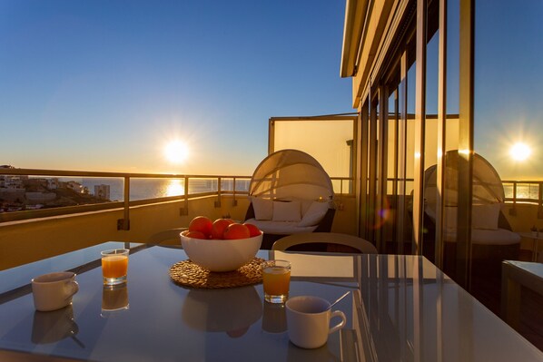 A balcony with a table set for breakfast, including a bowl of fruit, two glasses of orange juice, and a cup.