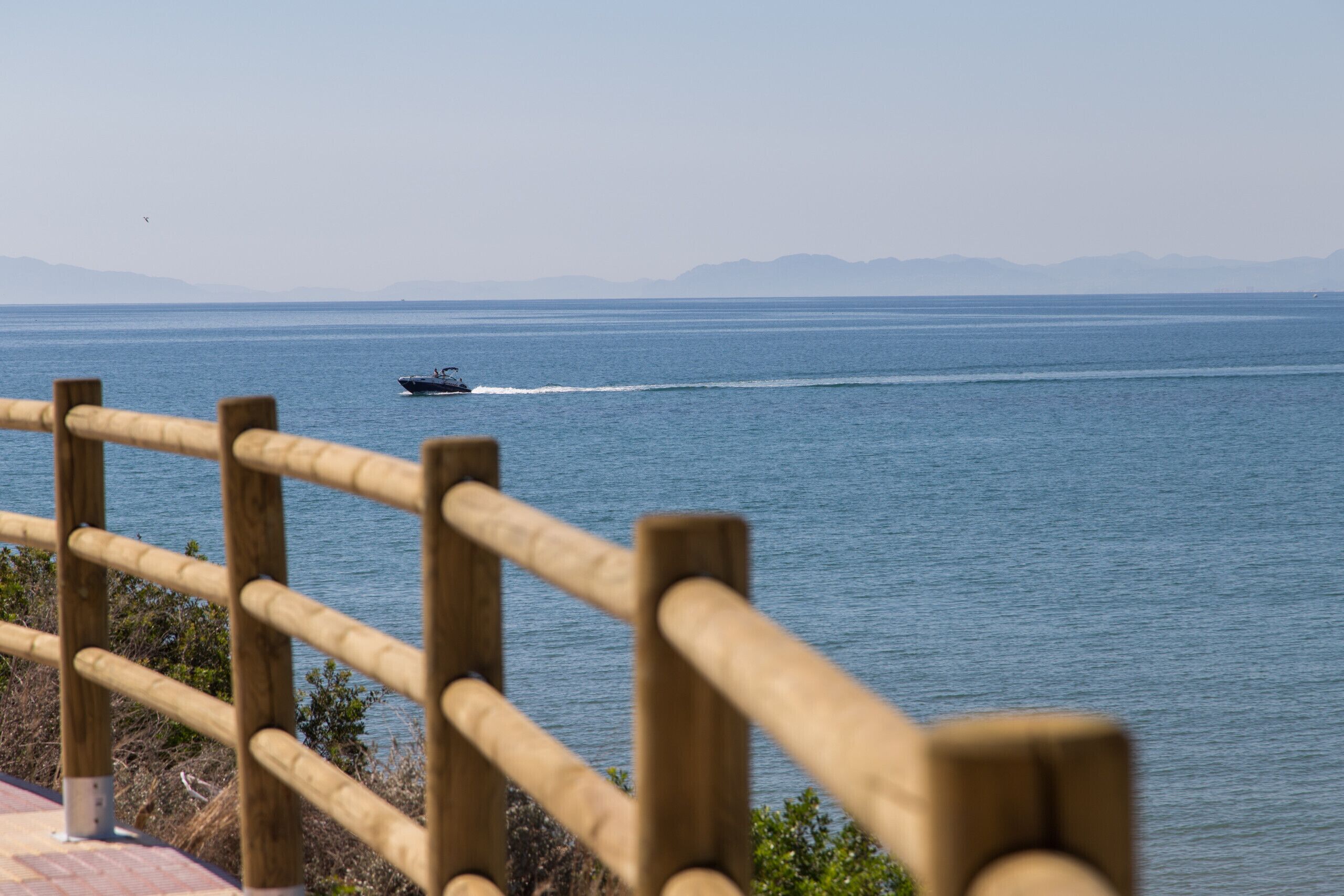 Eine Holzbrüstung mit Blick auf ein ruhiges Meer, in der Ferne ein Boot.