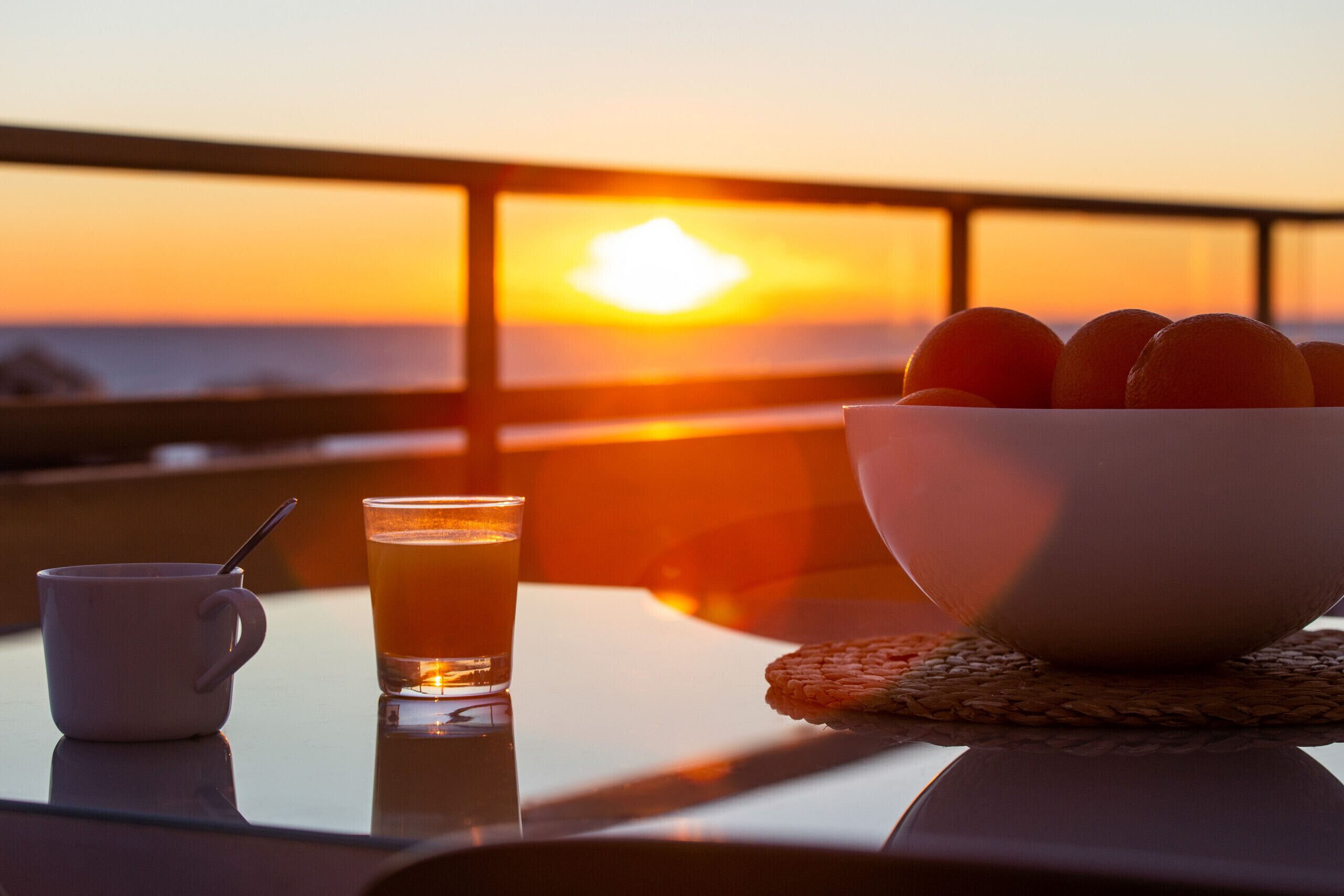 A table with a bowl of fruit, a glass of juice, and a cup, set against a sunset view.