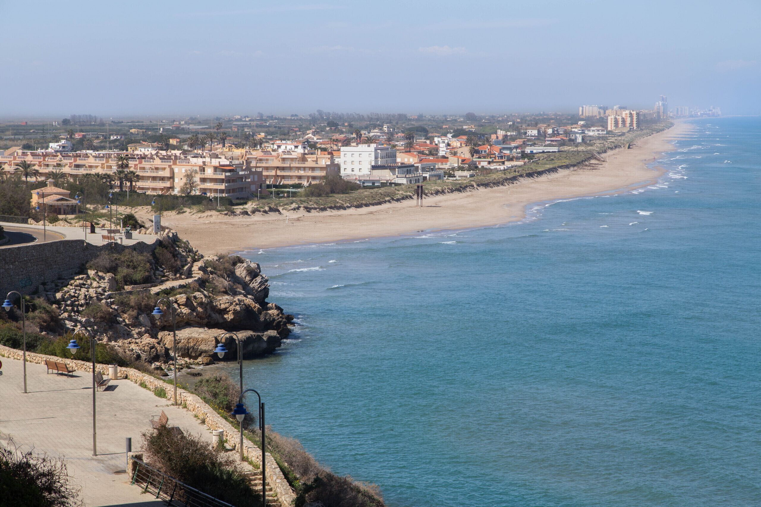 Un paisaje urbano costero con una playa de arena, costa rocosa y un paseo marítimo con bancos y farolas.