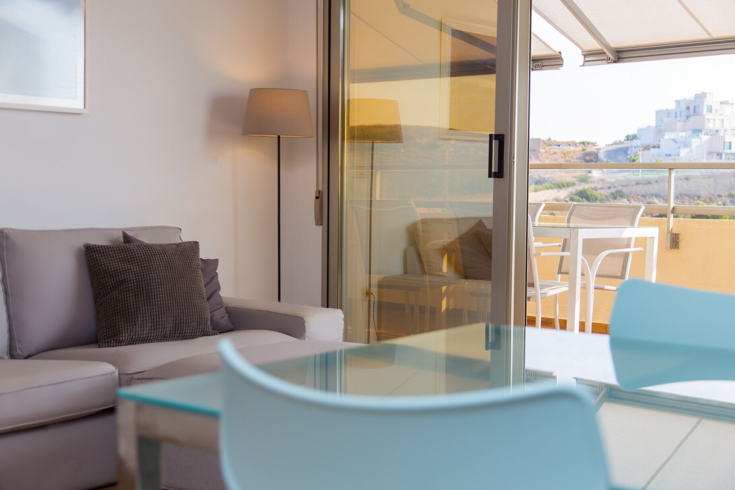 Un salon moderne avec une table en verre, un canapé et une vue sur un balcon aménagé avec des sièges d’extérieur.
