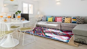 Un salon moderne avec une table à manger ronde, des chaises blanches, un canapé gris agrémenté de coussins colorés et un tapis aux motifs vifs.