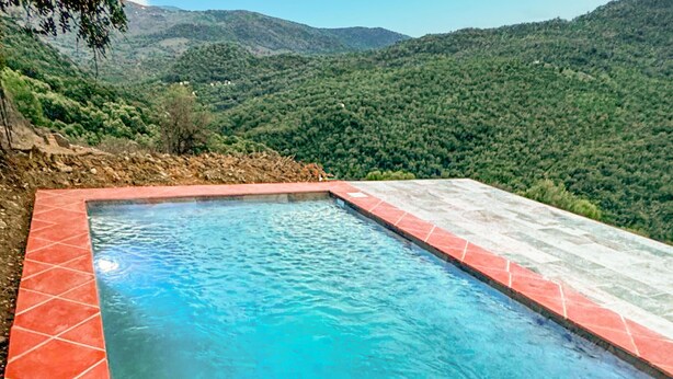 Una piscina rectangular con baldosas rojas y una superficie de agua azul cristalina, rodeada por una terraza de piedra, con colinas verdes exuberantes al fondo.