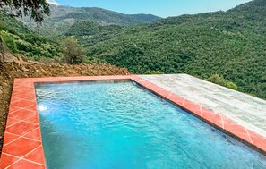 Una piscina rectangular con baldosas rojas y una superficie de agua azul cristalina, rodeada por una terraza de piedra, con colinas verdes exuberantes al fondo.