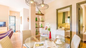 Habitación de hotel moderna con una mesa de comedor de vidrio, sillas blancas, un florero rojo con flores y un espejo grande.