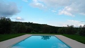 A rectangular swimming pool with a clear blue surface, surrounded by a light-colored deck, set against a backdrop of lush greenery and a partly cloudy sky. - Podere Lucignano Secondo Agriturismo (Gaiole in Chianti)
