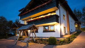 A two-story building with a balcony adorned with flowers, a wooden swing, and a well-lit entrance. - Branmontecrai (Bran)