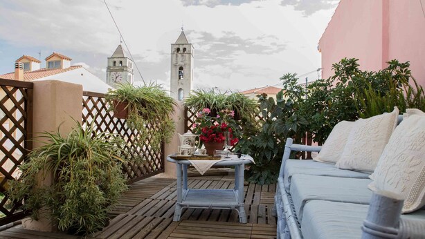 Una terraza de madera con un sofá de mimbre y una pequeña mesa con una tetera y flores. Se divisa una torre de campanas al fondo.