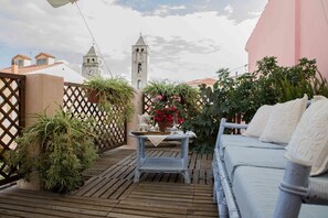 Una terraza de madera con un sofá de mimbre y una pequeña mesa con una tetera y flores. Se divisa una torre de campanas al fondo.