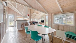 Un'ampia stanza con un grande soffitto in legno, un tavolo da pranzo centrale con sedie, un angolo cottura con sgabelli e un'ampia finestra che offre vista sull'esterno.