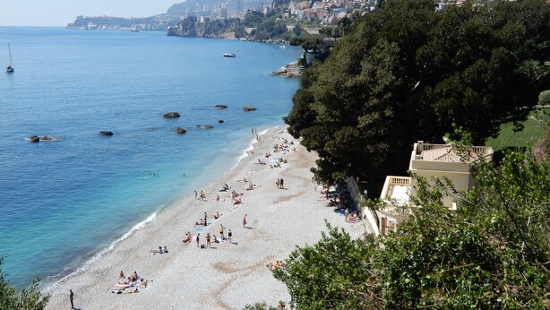 Una playa costera con aguas azules cristalinas, una orilla arenosa y una colina con edificios.