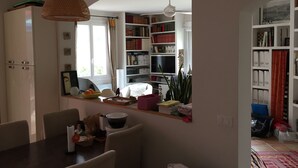 Un comedor con una mesa, sillas y una lámpara colgante. Se observan estanterías llenas de libros y un televisor al fondo.
