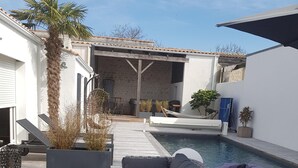 Uma área à beira da piscina com um sofá de vime, uma palmeira e um muro de pedra.