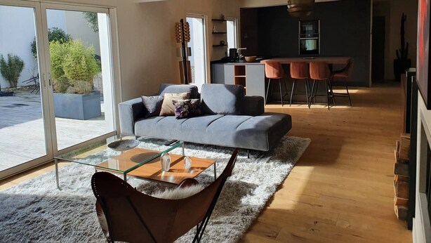 Uma sala de estar moderna com um sofá cinzento, uma mesa de centro de vidro e um tapete macio. Há uma área de jantar com um balcão e banquetas, e uma cozinha ao fundo.
