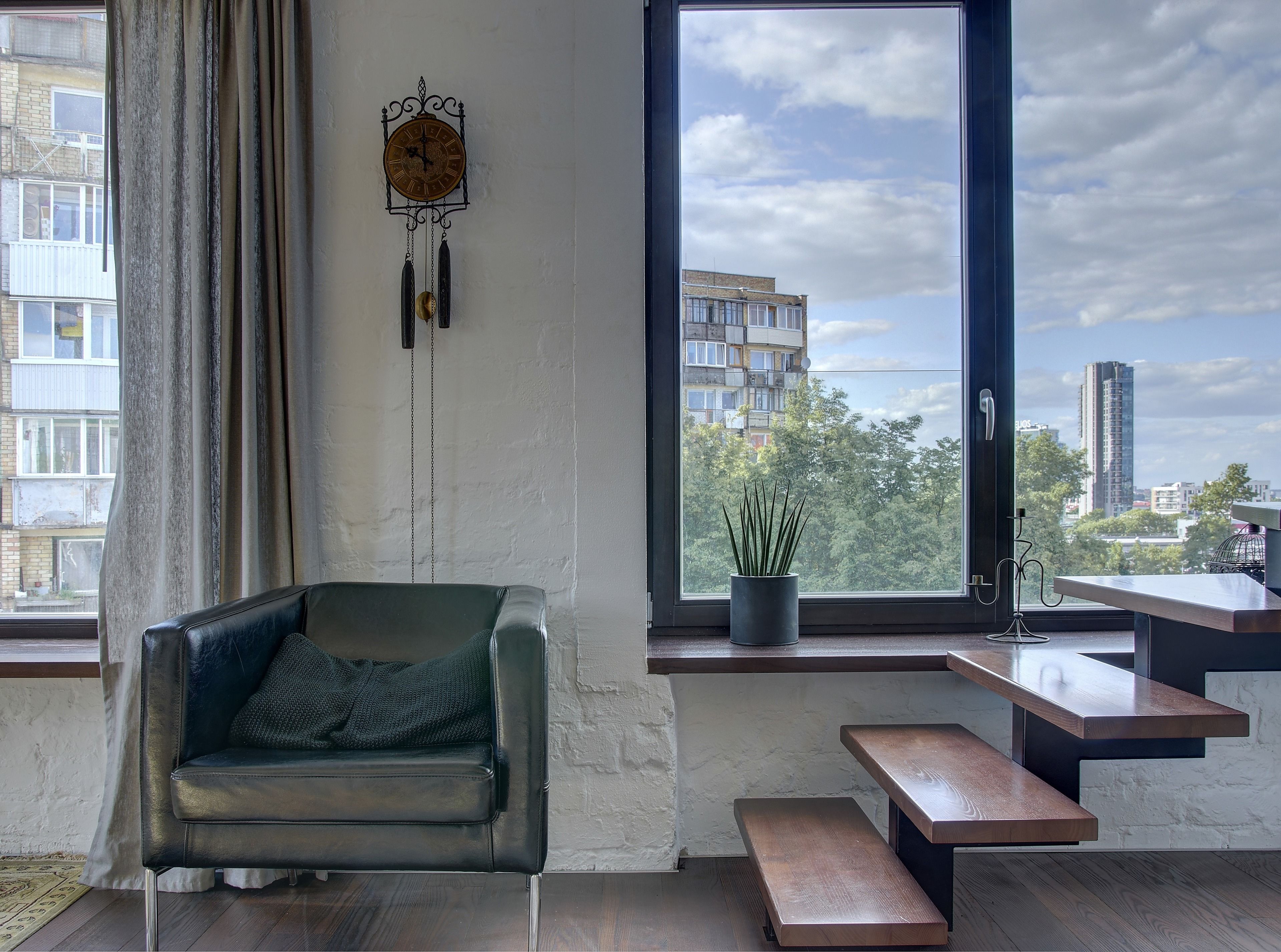 A modern living room with a leather sofa, a wooden bench, a potted plant, and a view of buildings through a large window.