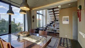 Ein moderner Essbereich mit einem Glastisch, Holzkästen, Pendelleuchten und Blick auf die Stadt durch große Fenster.