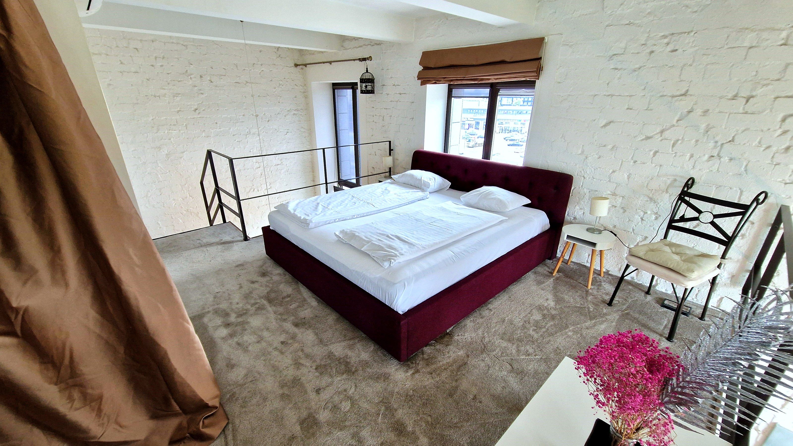 A bedroom with a large bed, a chair, a small table, and a window with a view of the street.