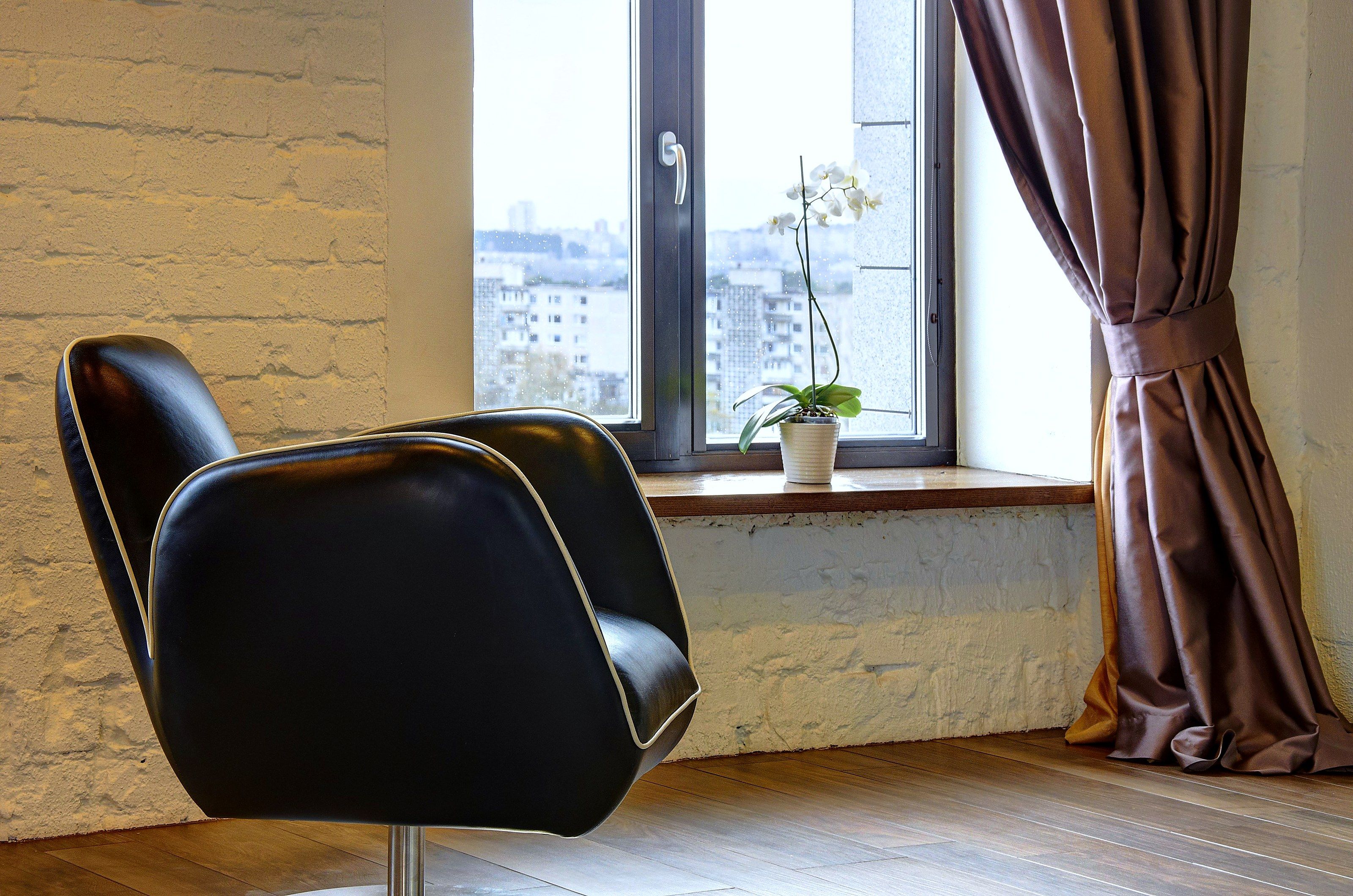 A room with a black leather chair, a window with a view of buildings, and a potted plant on the windowsill.