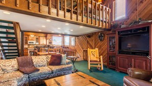 A cozy cabin interior with a wooden staircase, a dining area, a sofa, a coffee table, and a television.