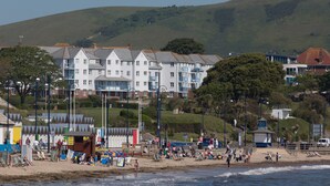 Un hotel frente a la playa con edificios de varios pisos, una playa de arena y una colina al fondo.