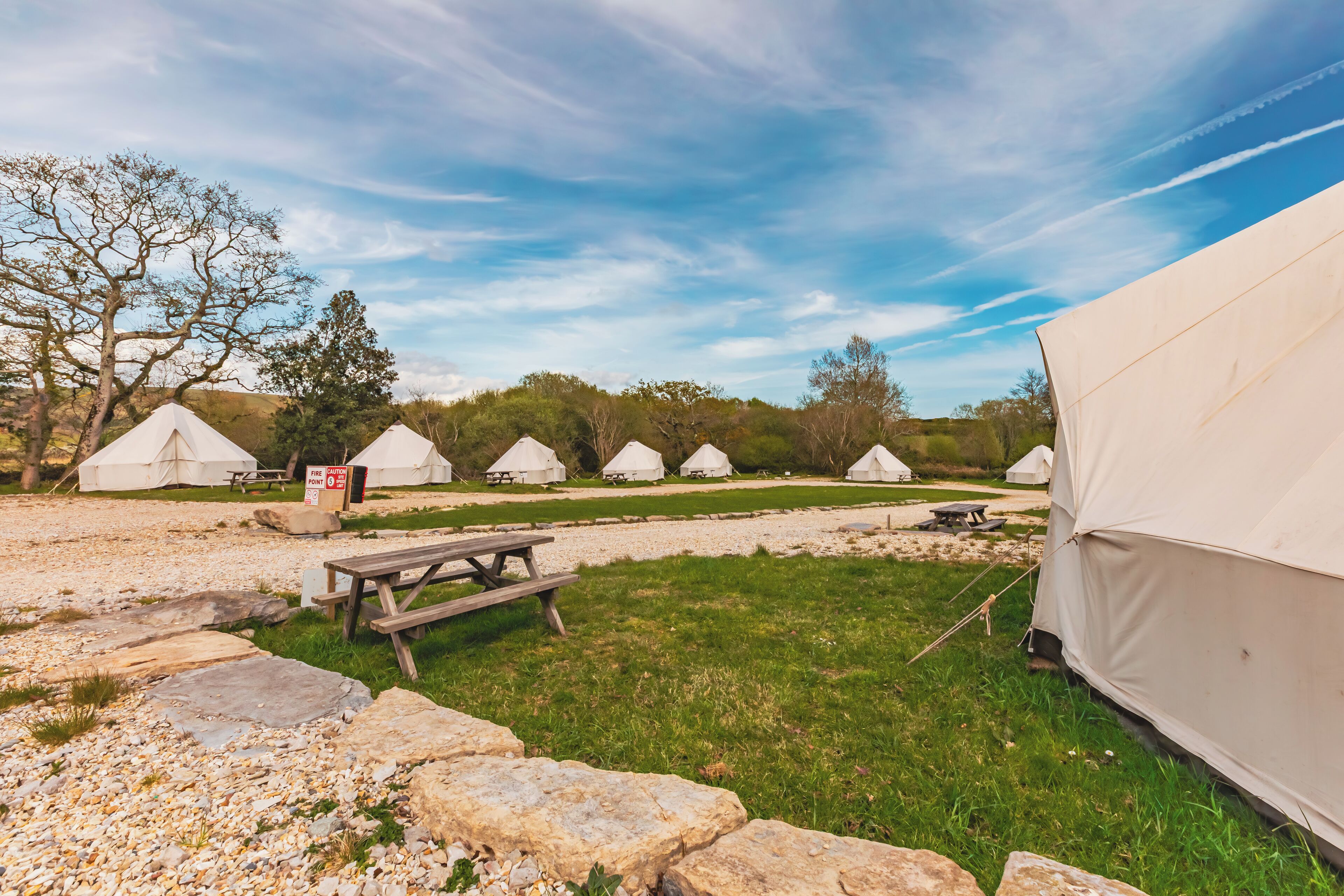 A row of white tents with picnic tables in a grassy area, surrounded by rocks and trees.