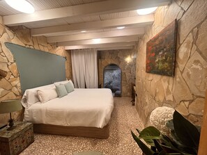 A bedroom with a stone wall, a bed with white linens, a nightstand with a lamp, and a decorative plant. - Boutique Hostal Salinas (Sant Josep de sa Talaia)