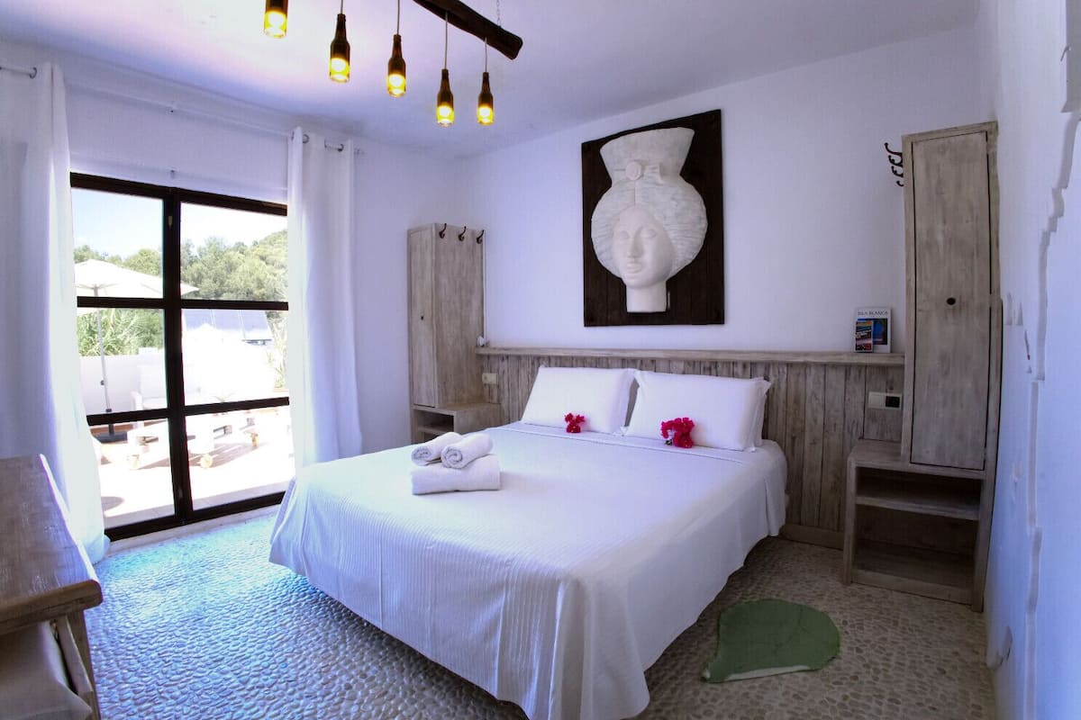 A bedroom with a large bed, a wooden headboard, a wall-mounted vase, and a view of the outdoors.