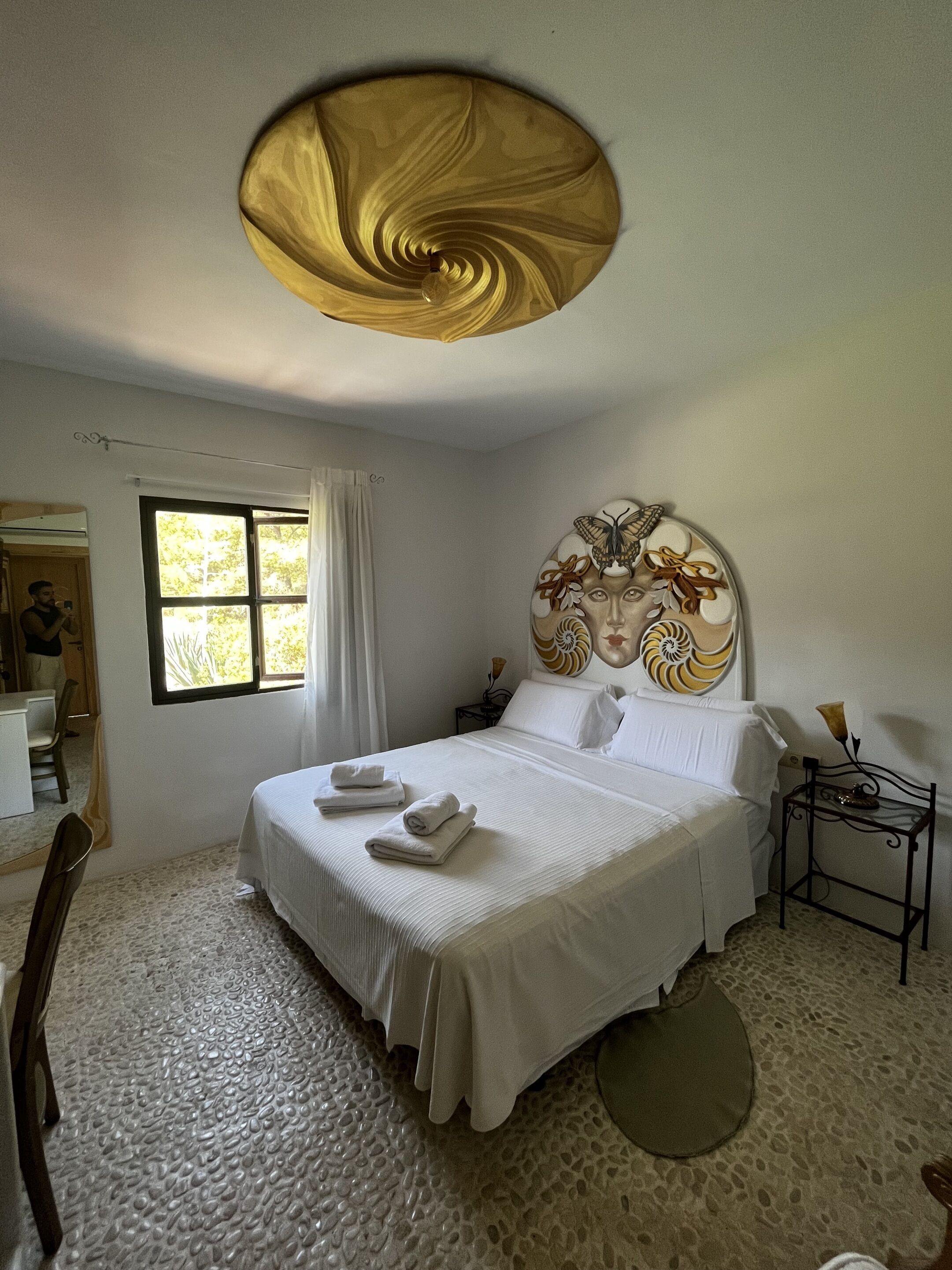 A hotel room with a large bed, a headboard with a decorative design, a bedside table with a lamp, and a window with curtains.