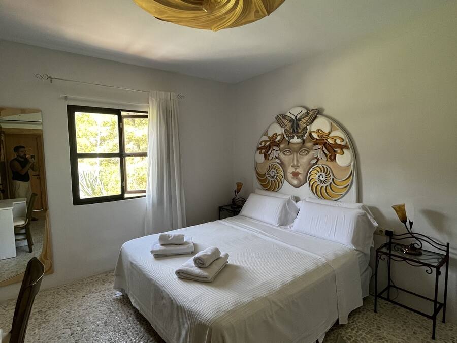 A hotel room with a large bed, a headboard with a decorative design, a bedside table with a lamp, and a window with curtains.