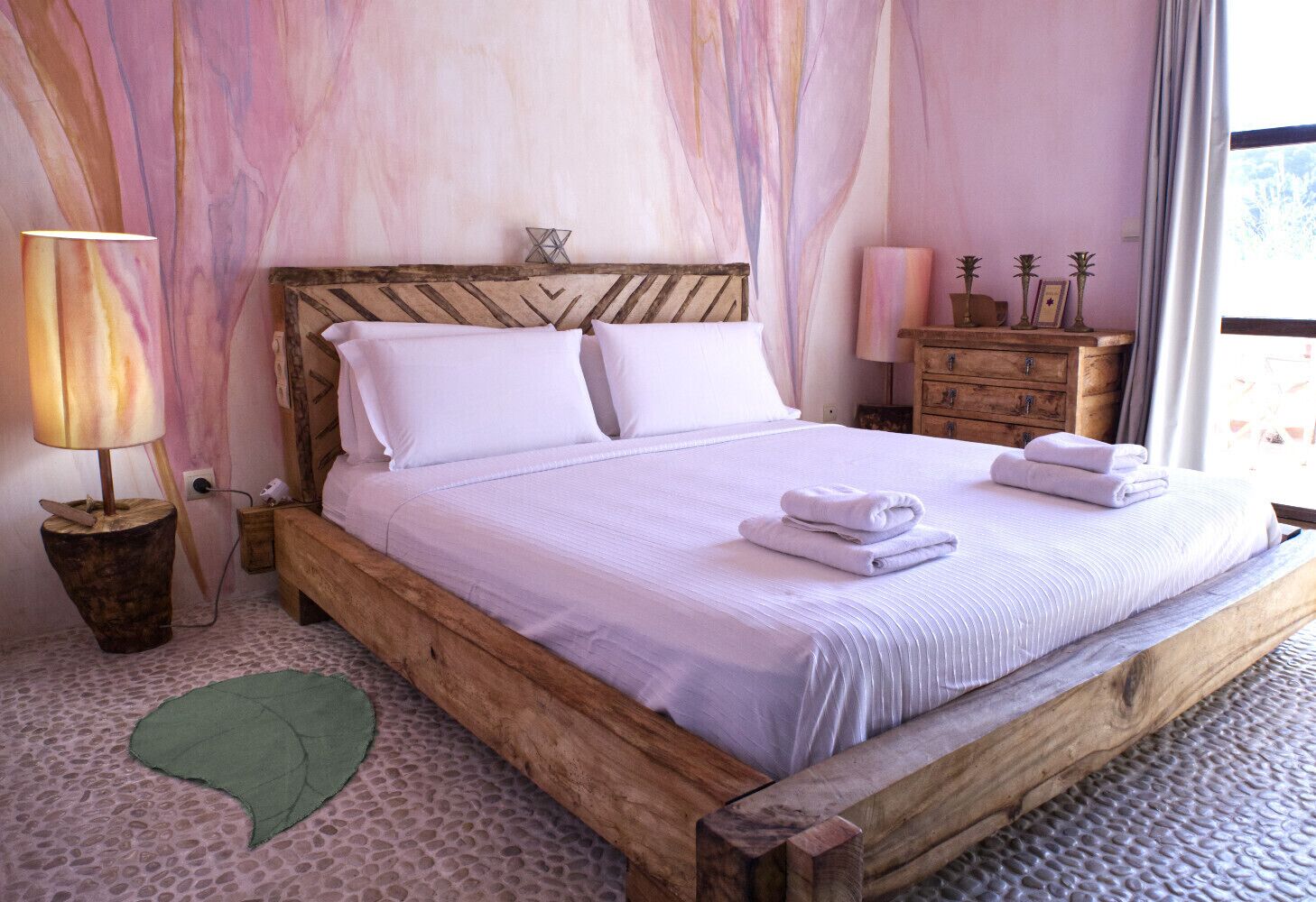 Een houten bed met witte lakens en kussens, een bedlamp, een nachtkastje van hout en een groen decoratief object op de vloer.