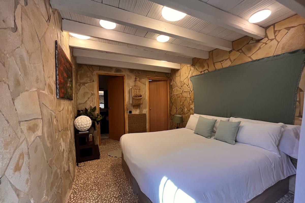 A bedroom with a stone wall, a bed with white bedding, a stone floor, and a ceiling with recessed lighting.